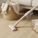 A room is being cleaned. A pink handheld vacuum cleaner is positioned diagonally across the image, moving from the lower left to the lower center. The floor is light oak parquet, with a herringbone pattern. A light beige woven basket with cream-colored tassels sits near the center left. A cream-colored throw blanket or knitted sweater is on top of the basket. A light beige couch with a cushioned seat is in the upper right. A light gold wire basket, containing cleaning spray bottles and cloths, is placed on the floor near the bottom right. A light brown wicker armchair is located in the upper left corner. Natural light illuminates the scene, creating a bright and clean atmosphere. The overall style is minimalist and modern. The focus is on the practical aspects of home cleaning.