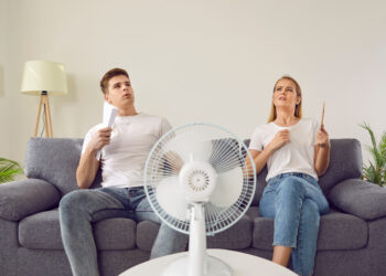 Young couple suffering from hot summer weather and sitting on sofa with electric fan