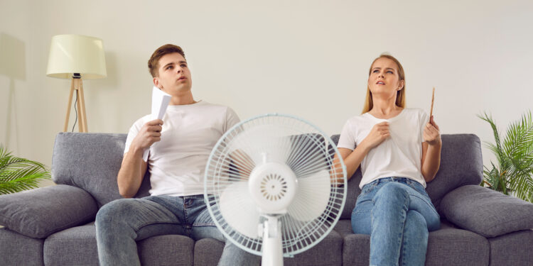 Young couple suffering from hot summer weather and sitting on sofa with electric fan