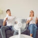 Young couple suffering from hot summer weather and sitting on sofa with electric fan